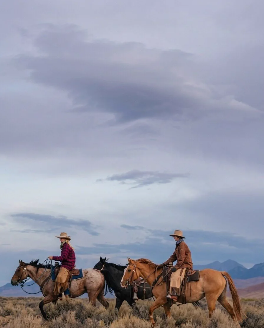 Two cowboys riding horses through an open Idaho desert landscape under a cloudy sky